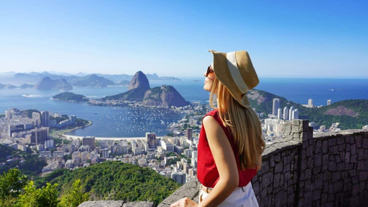 A woman wearing a straw hat and sunglasses is standing on a viewpoint, overlooking a city and coastal landscape with mountains and water in the background, enjoying the tranquility of female solo travel.