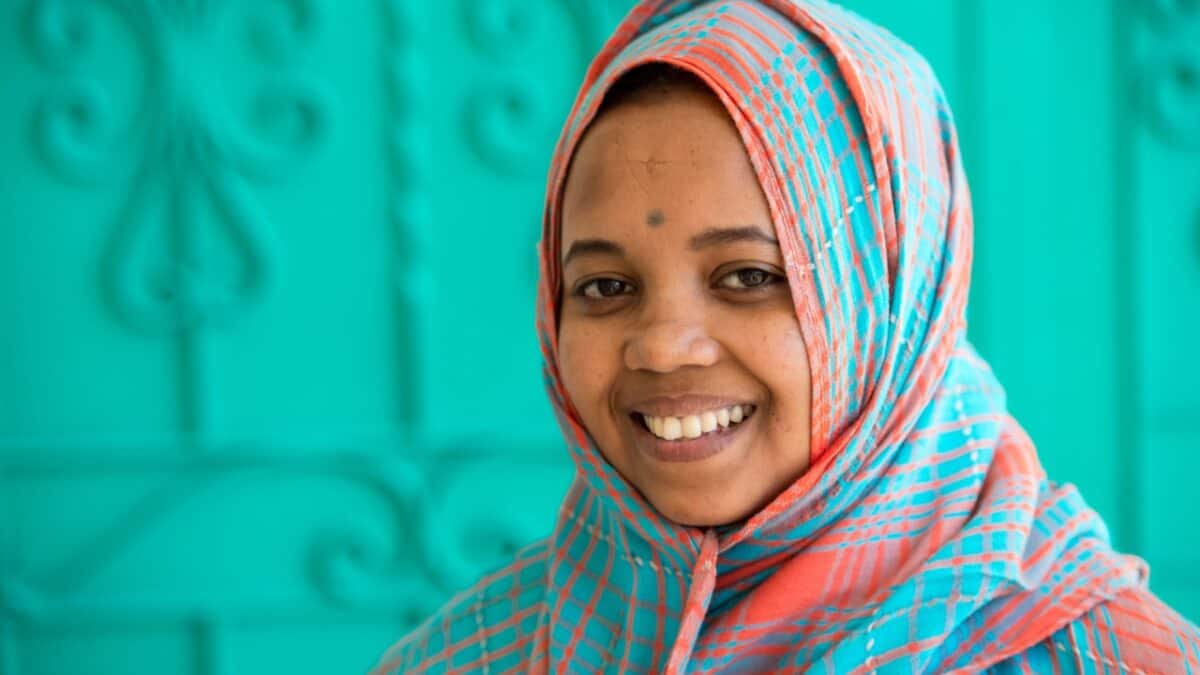 A woman wearing a brightly colored headscarf smiles at the camera, standing in front of a turquoise background, embodying the spirit of female solo travel.