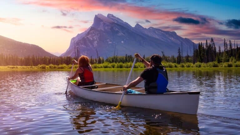 Two people paddle a canoe on a calm lake with a mountain and sunset in the background.