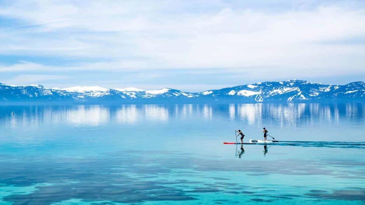 15 Best Cities In The United States For Thanksgiving Celebrations 10 Two people paddleboarding on a clear, calm lake with snow-capped mountains in the background under a blue sky.