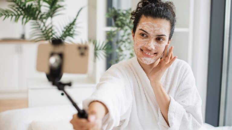 Person in a white robe taking a selfie with a smartphone, wearing a facial mask, smiling, and touching their cheek.