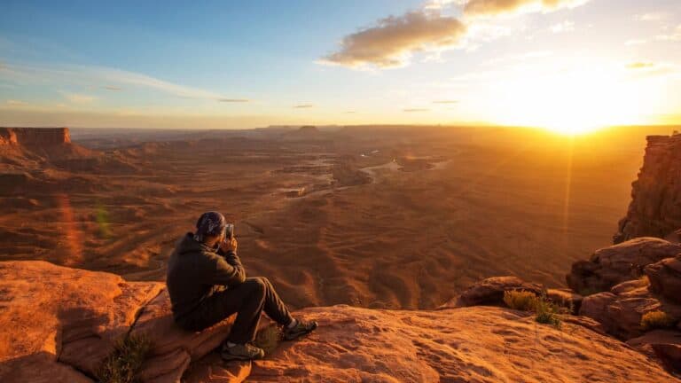 Person sitting on a rocky ledge, photographing a vast canyon landscape at sunset with a camera.