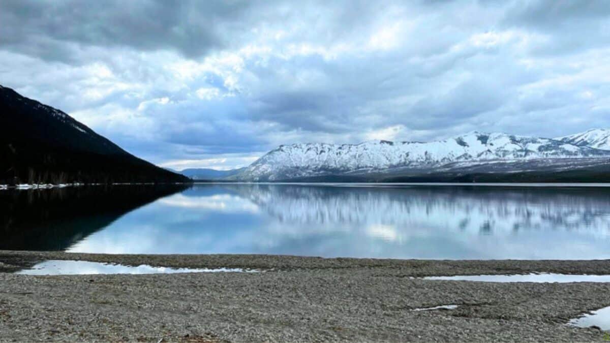 Serene lake with snowy mountains in the background, under a cloudy sky, reflecting calm water and a sandy shore.