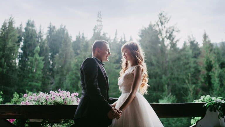 A couple in wedding attire hold hands and smile at each other on a balcony with a forest background.
