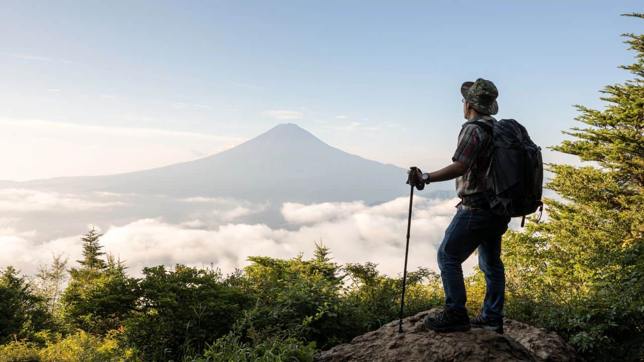 A hiker stands on a rocky outcrop, gazing at a distant mountain peak, surrounded by lush greenery and clouds.