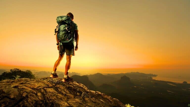 A person with a large backpack stands on a rocky cliff, overlooking a scenic sunrise or sunset view of mountains and valleys.