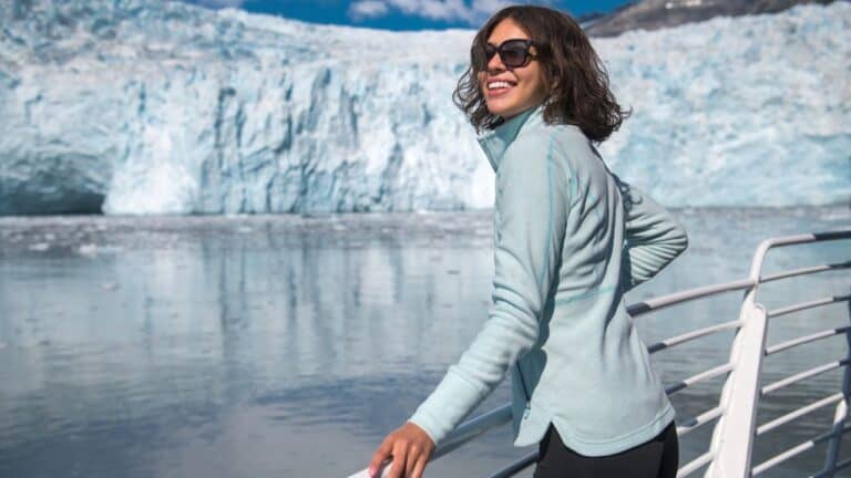 A person in a light jacket smiles while leaning on a railing with a large glacier in the background.