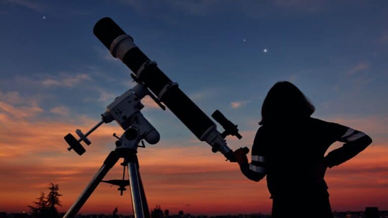 Silhouette of a person looking through a telescope at sunset with a bright star visible in the sky.