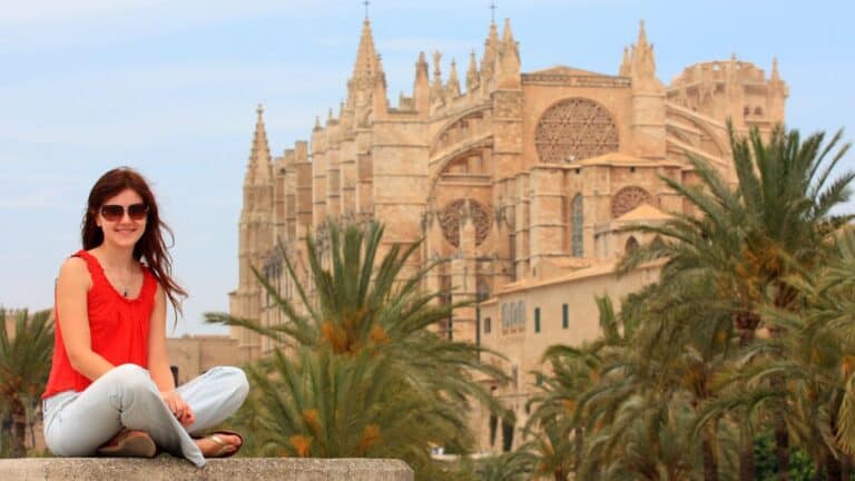 Smiling woman in sunglasses and red top sits on ledge with a grand cathedral and palm trees in the background.