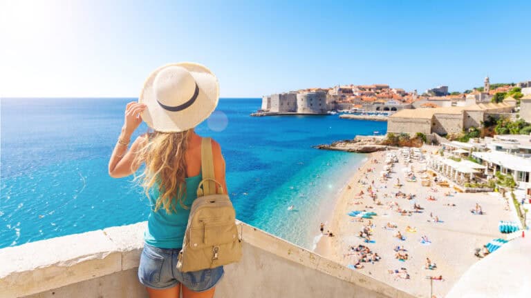 A woman in a hat and backpack looks out over a scenic coastal town with a beach and clear blue water.