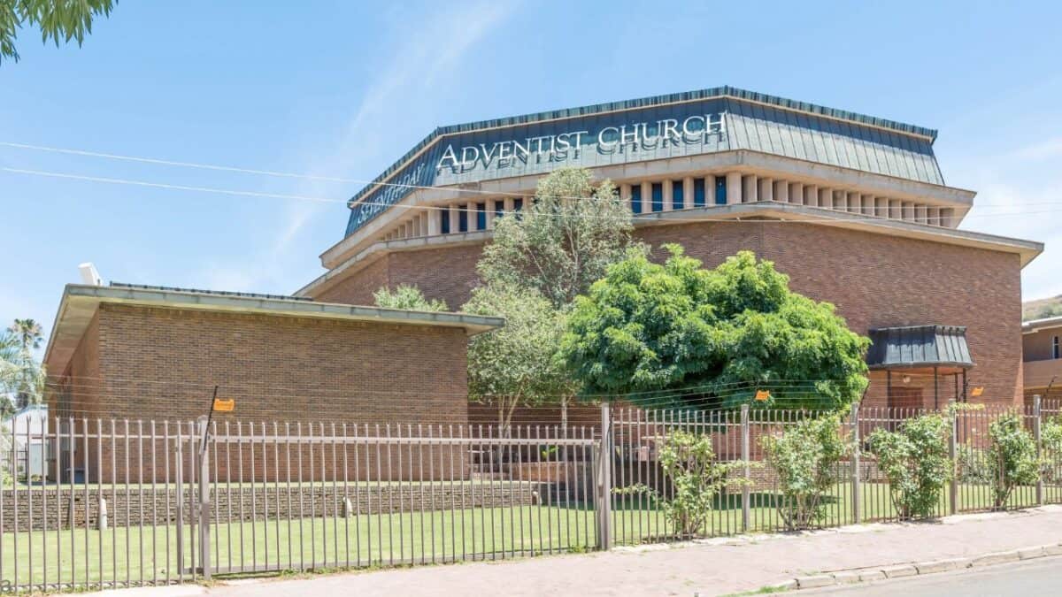 Exterior of a large brick Adventist church with a green fence and trees in the foreground.