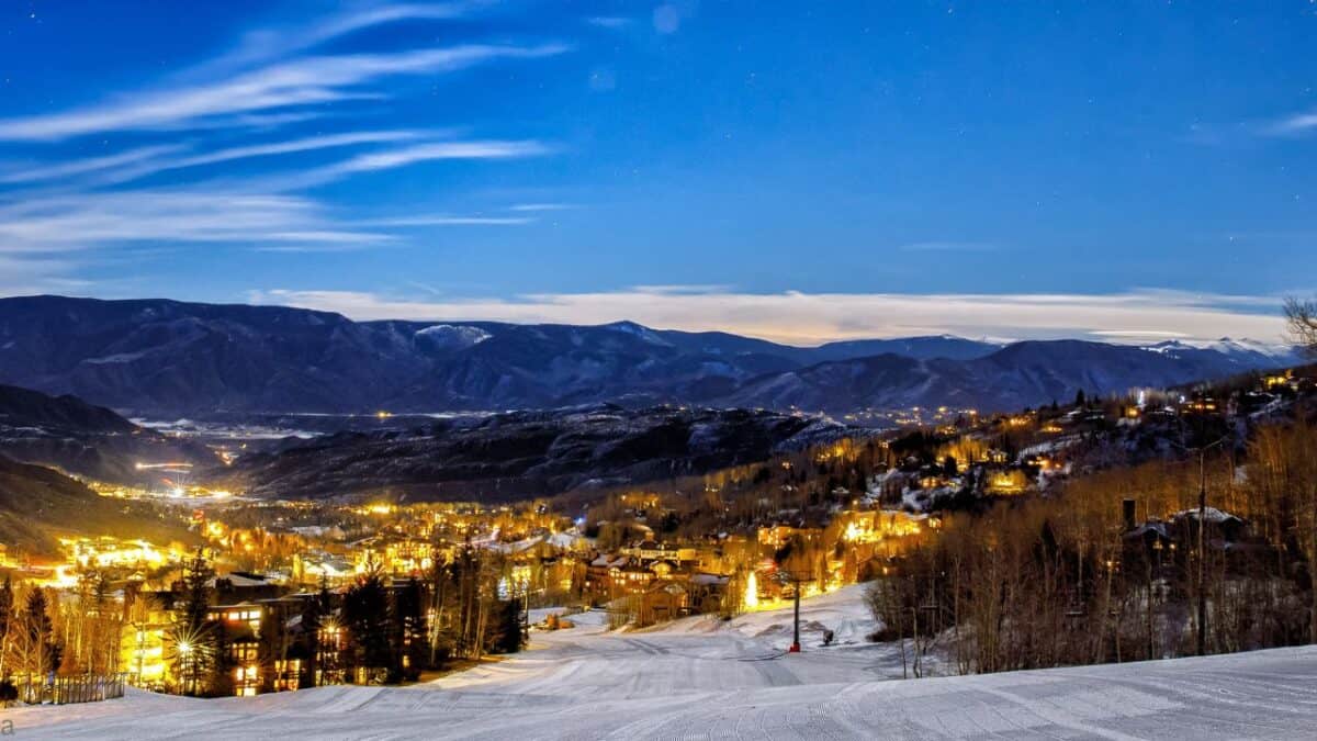 Snowy mountain village illuminated at night, under a starry sky and surrounded by hills.