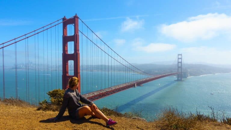 A woman in a striped dress and pink shoes sits on a grassy hill overlooking the Golden Gate Bridge in San Francisco.