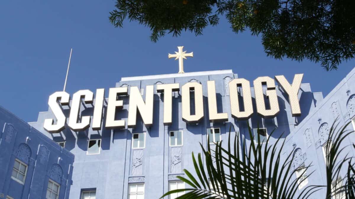 The Church of Scientology blue building with a prominent rooftop sign and a cross, framed by trees.
