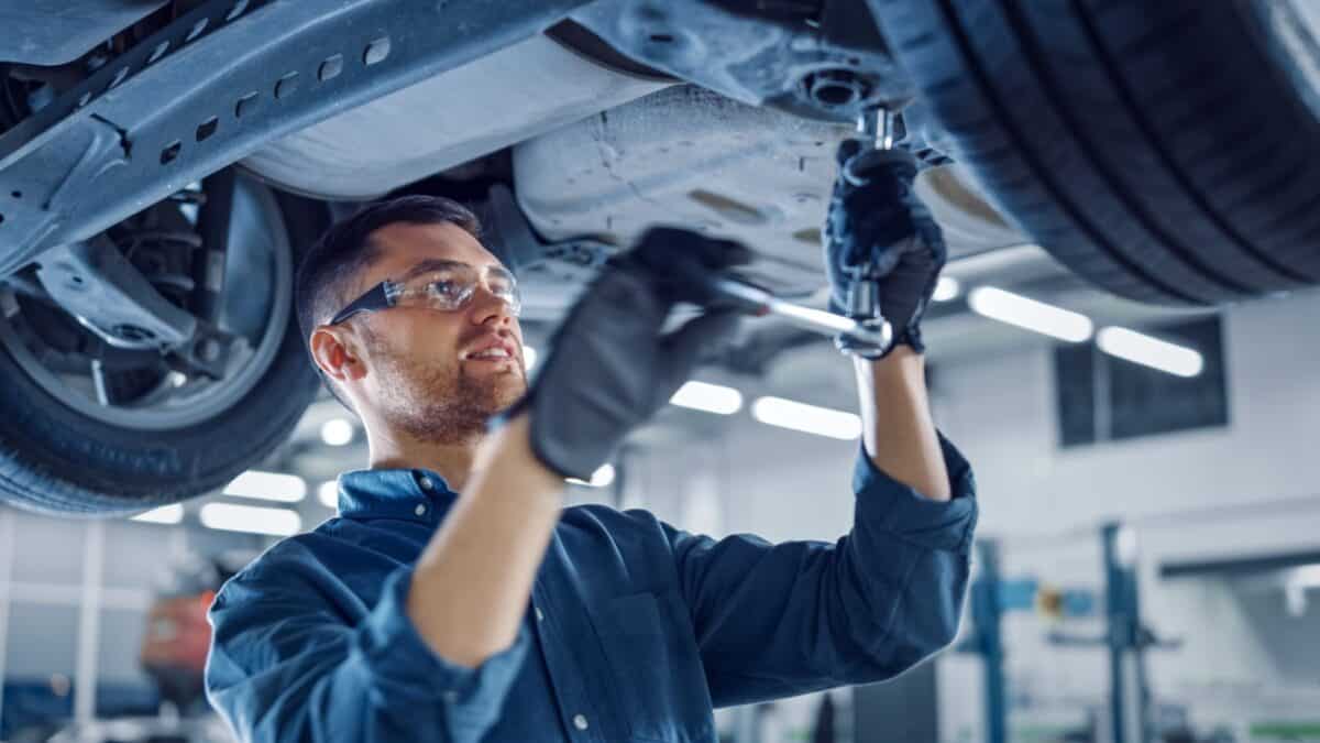 A mechanic wearing safety glasses and gloves works under a car in a well-lit garage.