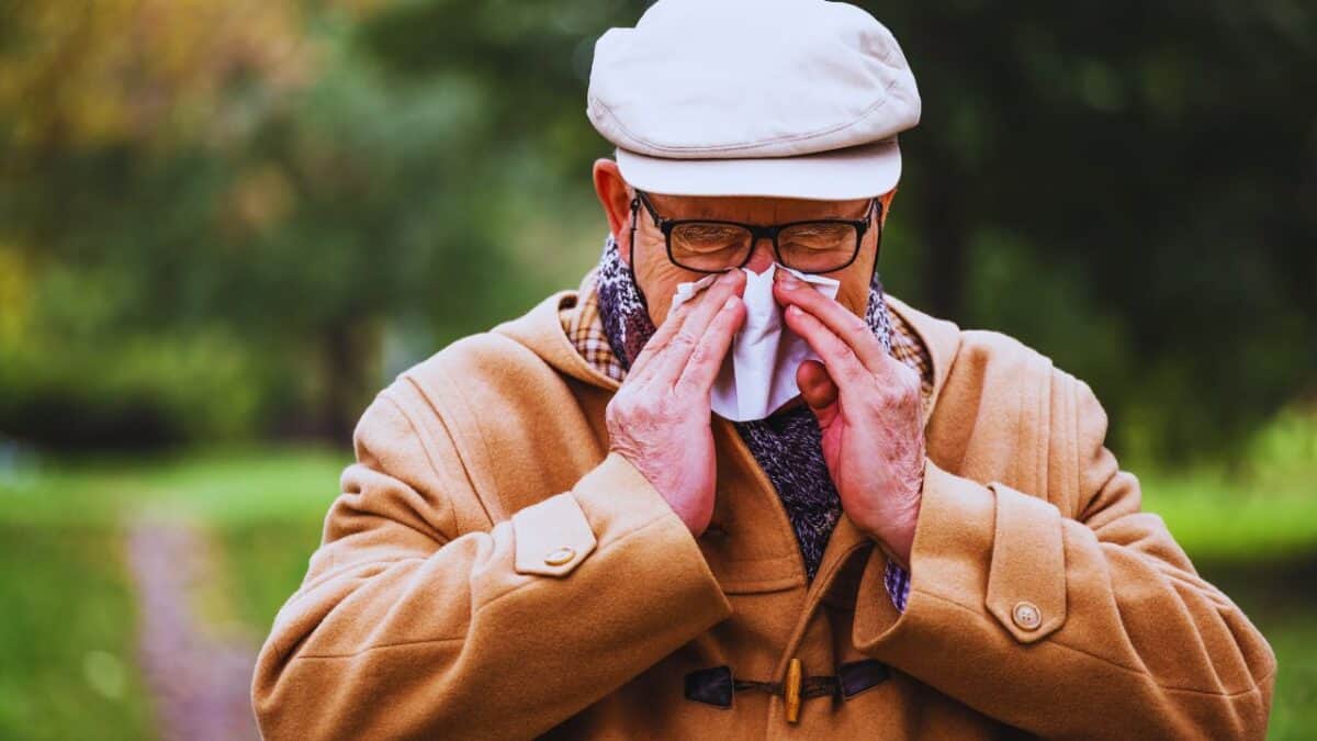 An elderly man in a coat and hat sneezes into a tissue outdoors in a park.