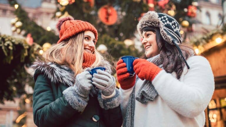 Two women in winter clothes smile and hold mugs in front of a decorated Christmas tree.