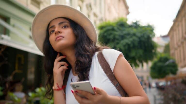 Woman in a hat holding a smartphone, looking up while strolling through a city street.