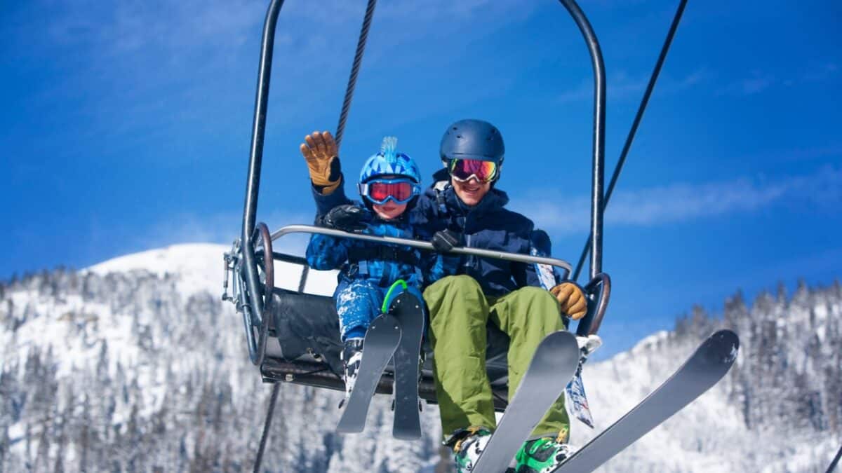 Two people on a ski lift in snowy mountains, wearing helmets and ski gear, smiling and waving.