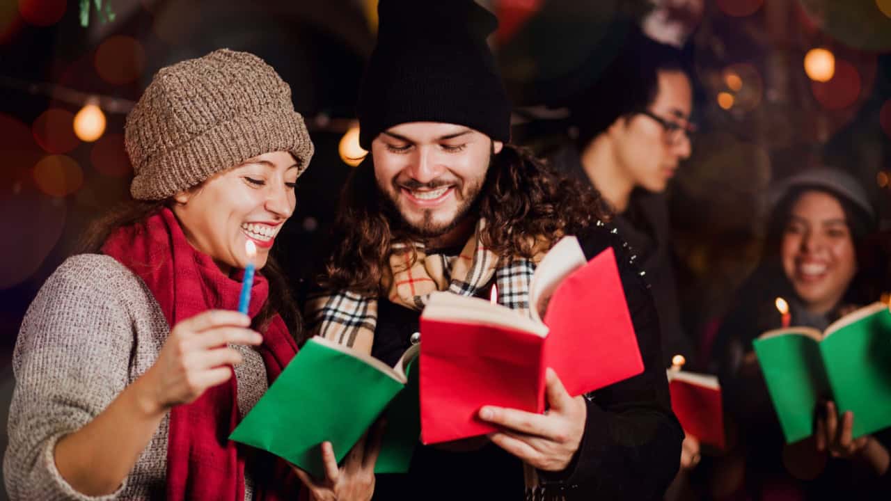 Group of people singing outdoors, wearing winter clothes, and holding songbooks and candles at night.