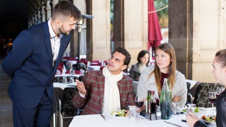 Group dining outdoors, man in checkered jacket talking to waiter, others listening.