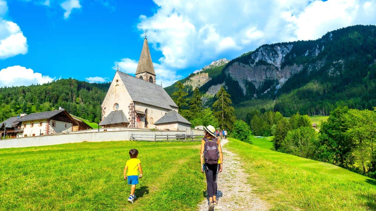 A woman and child walk on a path toward a church in a green, mountainous landscape under a blue sky.