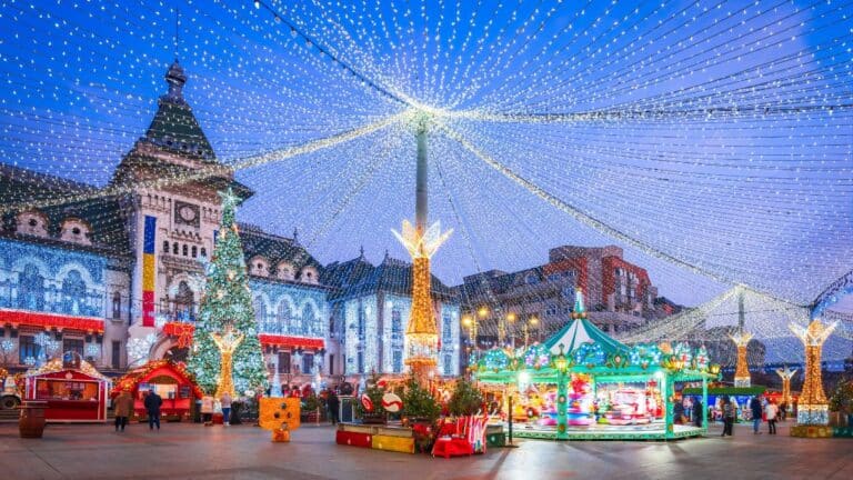 Festive town square with a carousel, Christmas lights, and decorated trees in front of historical buildings at dusk.
