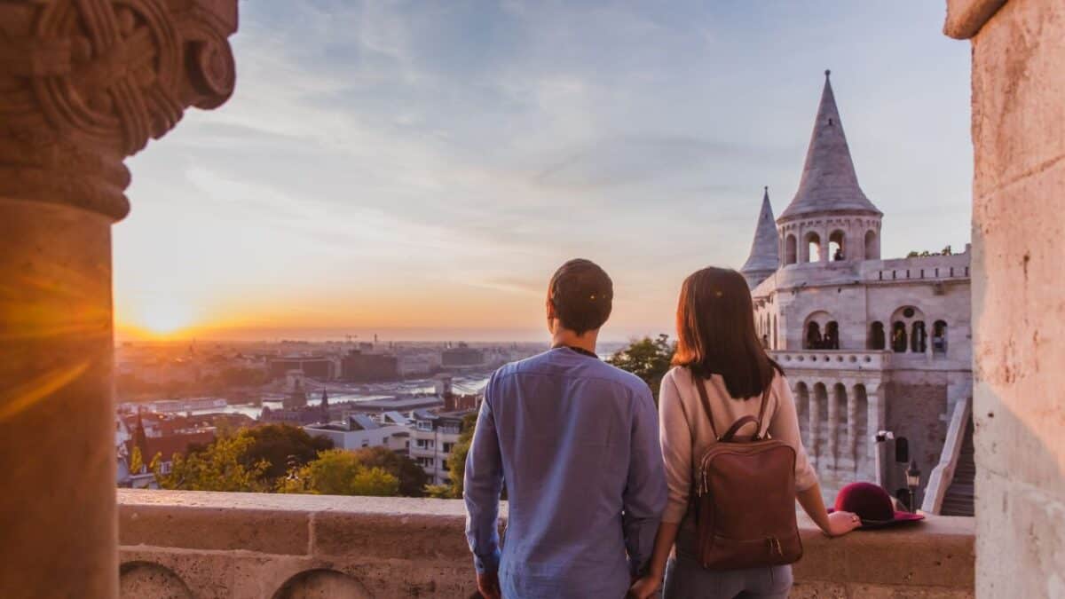 A couple enjoys a sunset view of a cityscape from a historic balcony, featuring tall spires and stone architecture.