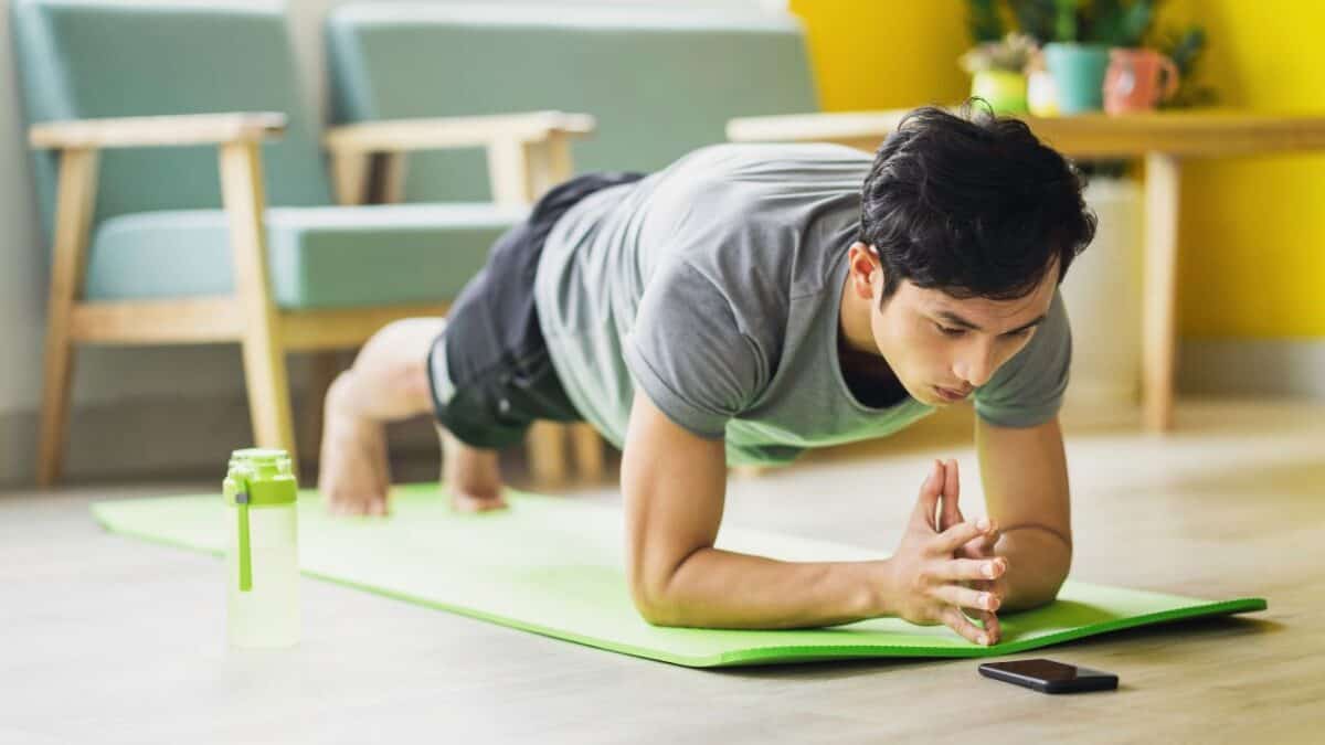 Person planking on a green yoga mat, looking at a smartphone. Water bottle nearby, bright room.