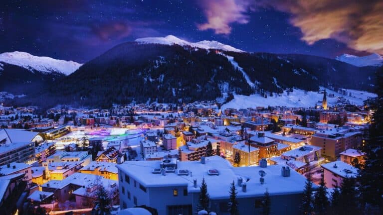 Snow-covered town at night with illuminated buildings, surrounded by mountains, under a starry sky.