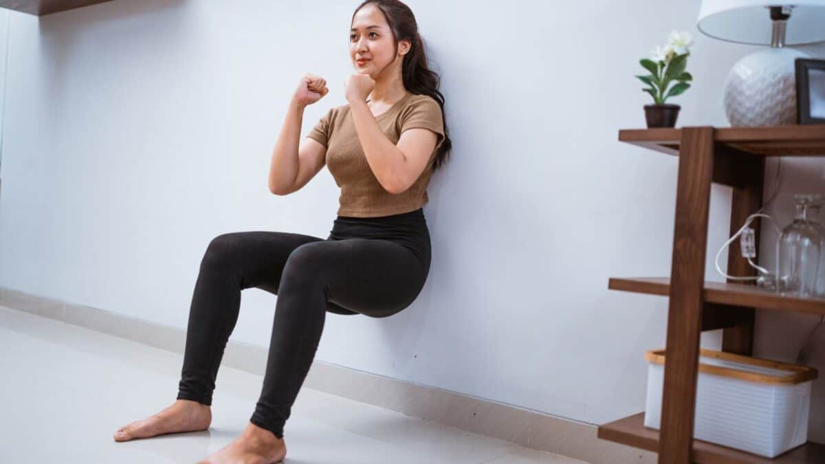 A person is doing a wall sit exercise indoors next to a shelf with plants and decorative items.