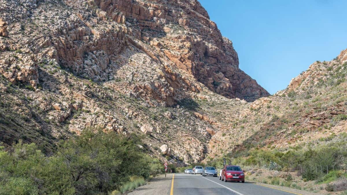 A scenic view of cars driving along a winding road through rugged rocky mountain terrain. The road curves around steep cliffs, with tall pine trees scattered across the landscape. Snow-capped peaks rise in the distance, and the sky is clear, with a hint of sunlight illuminating the rocky slopes. The vehicles are traveling at different speeds, with some closer to the foreground and others farther along the road, set against the dramatic, mountainous backdrop.