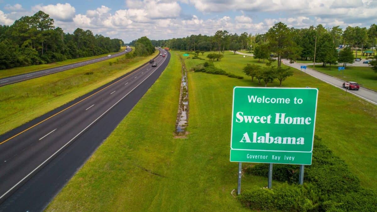 Roadside sign reading 'Welcome to Sweet Home Alabama' with a reference to Governor Kay Ivey. The sign is located along a well-maintained highway bordered by green grass and trees, with light traffic under a partly cloudy sky.