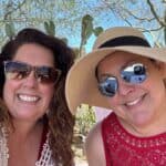 Two women smiling outdoors, both wearing sunglasses; one in a sun hat, with cactus plants in the background.