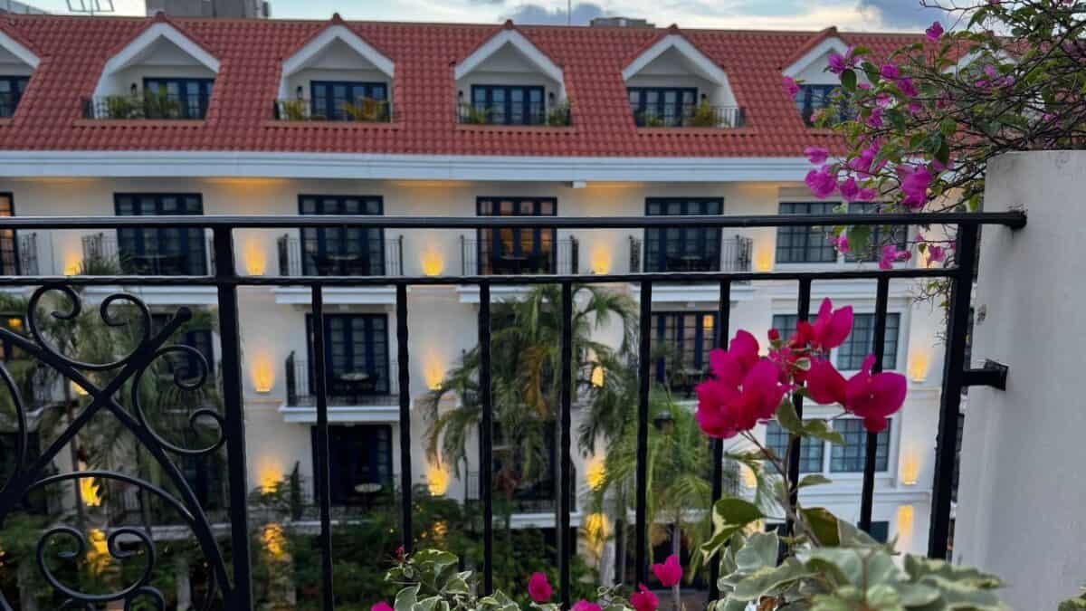 Pink flowers on a balcony overlook a white building with red roof and black windows, surrounded by palm trees.