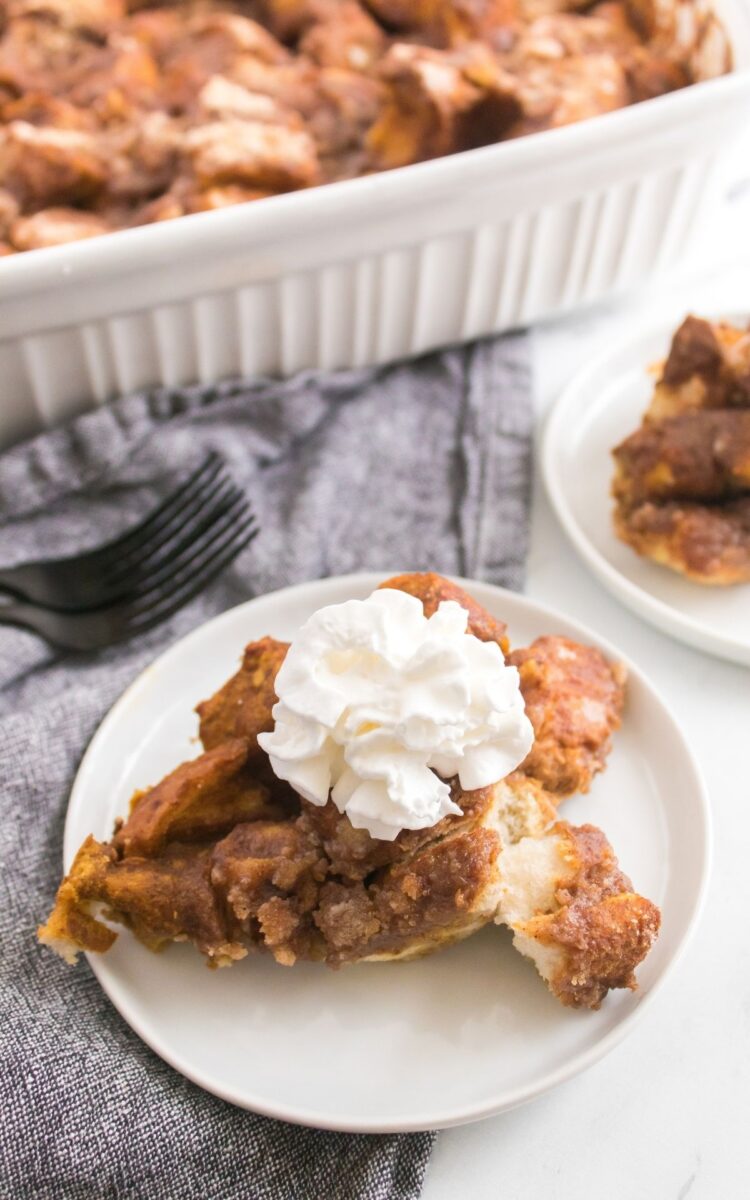 A slice of bread pudding topped with whipped cream on a white plate, with a casserole dish in the background.