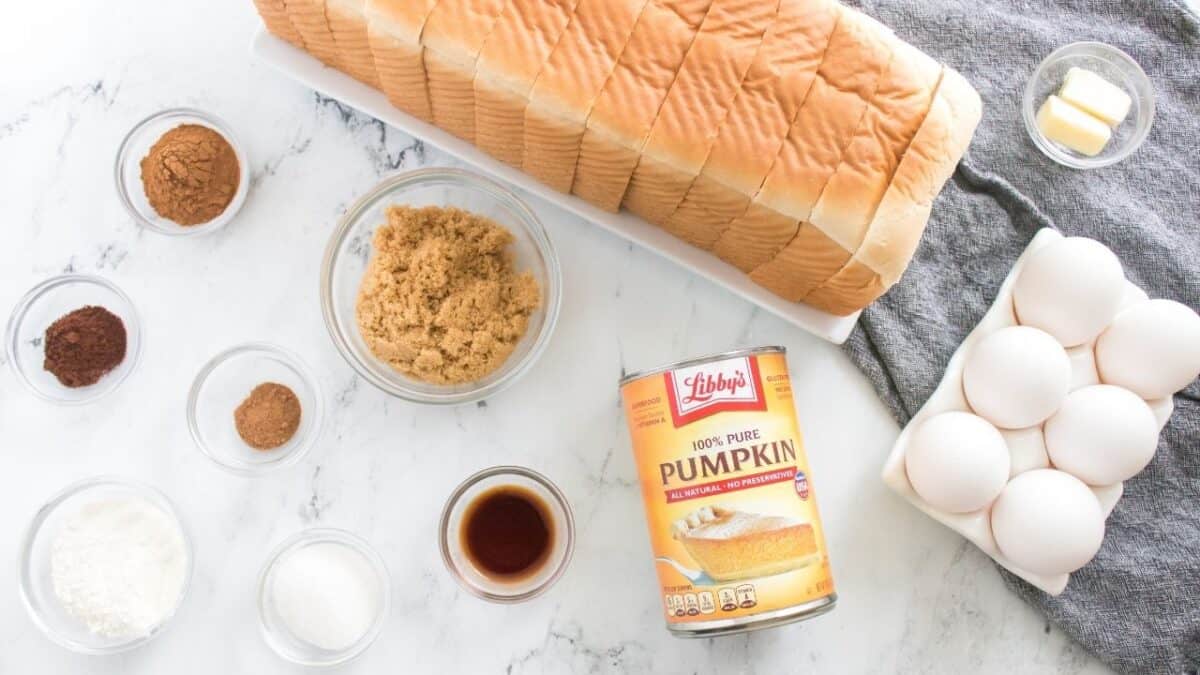 Sliced bread, eggs, canned pumpkin, spices, brown sugar, butter, vanilla, and flour on a white surface.