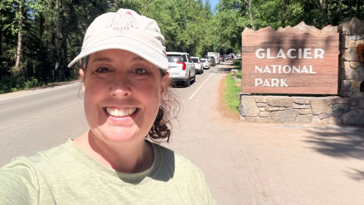 A smiling woman taking a selfie next to a Glacier National Park entrance sign with a line of cars on the road.
