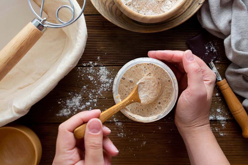 Hands holding a wooden spoon stirring sourdough starter in a glass jar on a wooden table.