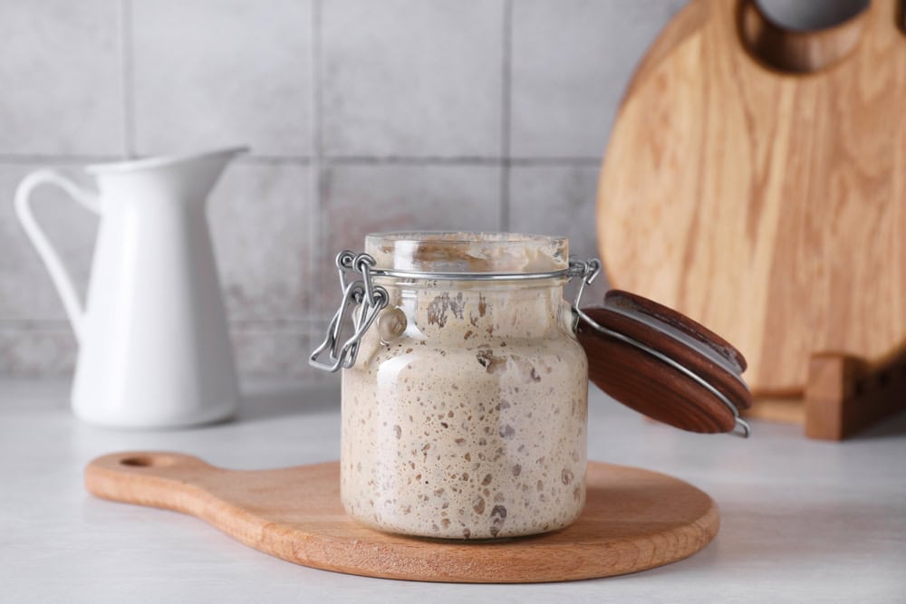 A glass jar filled with sourdough starter sits on a wooden cutting board.