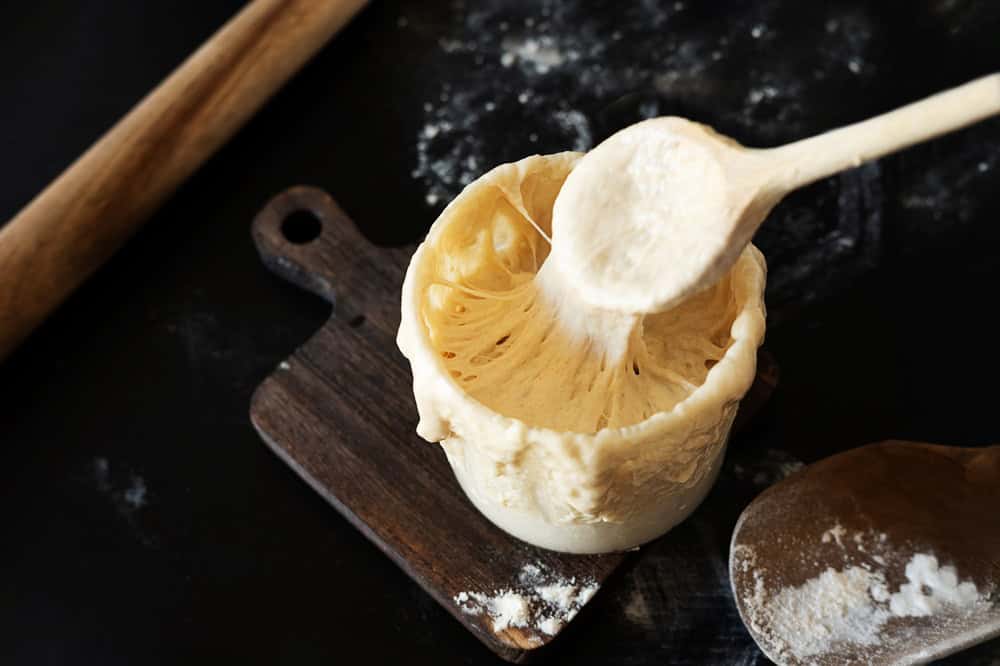 A wooden spoon lifts stretchy sourdough starter from a jar on a cutting board.